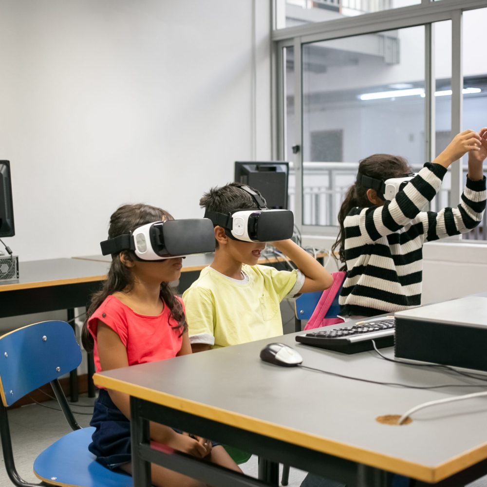 Cute multiethnic kids learning to use virtual reality glasses. Concentrated children sitting at table together and wearing VR headsets in classroom. Informatics, study and education concept